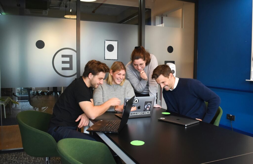 A group of four people enthusiastically collaborating and discussing blockchain technology around a laptop in a modern office.